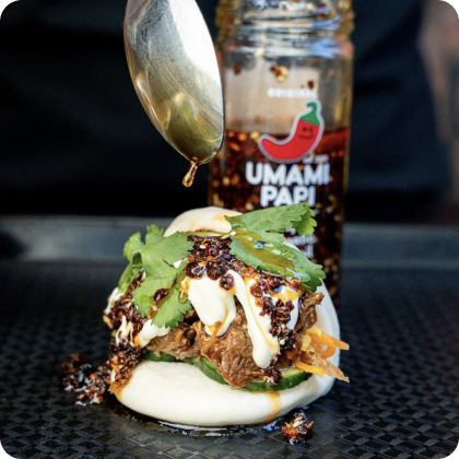 UmamiPapi recipes link. features Bao bun with pulled pork, cucumber & coriander with UmamiPapi Chilli Oil being dripped over the bun. Jar of Chilli Oil sits in the background. 